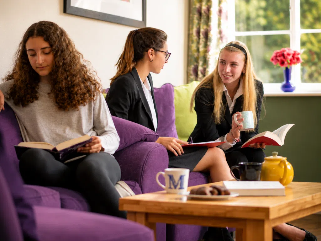 Three young women sit on a purple sofa, reading and chatting in a bright living room with mugs and a teapot on a wooden table.