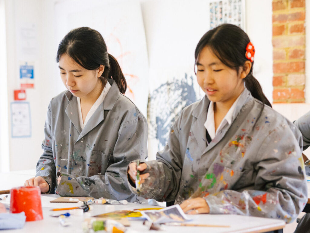 Two young girls in paint-splattered smocks painting at a table in a bright art studio.